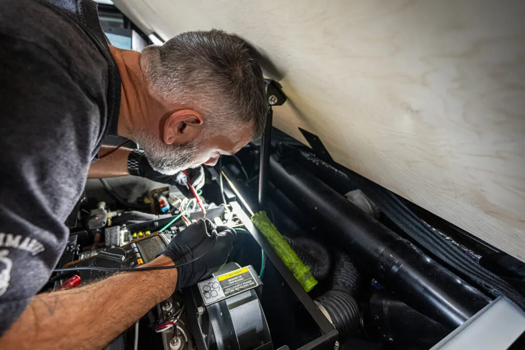 An experienced technician with short hair and beard working intently inside a semi-truck, repairing electrical components. He is wearing gloves, surrounded by wires and tools.
